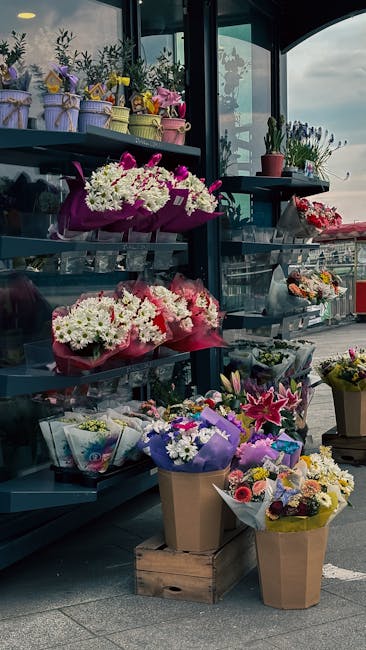 Display of various flower arrangements and bouquets at a flower shop, featuring white chrysanthemums and pink roses wrapped in pink and lavender tissue paper, placed on shelves and in brown paper wrap