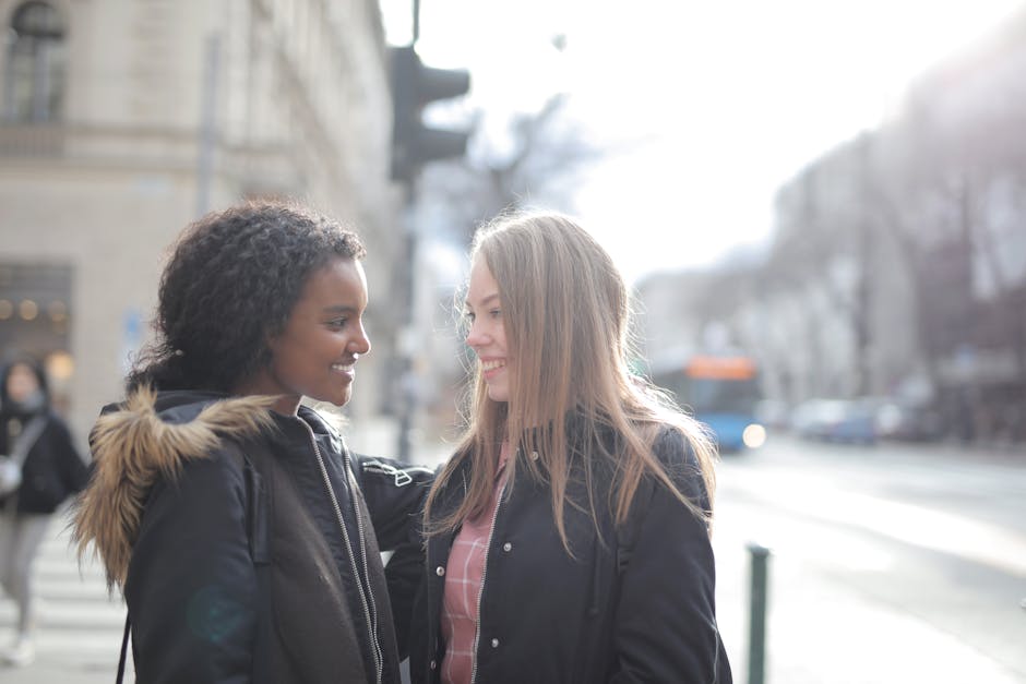 Two young women exchanging a friendly smile on a city street during daytime. One woman has dark, curly hair and is wearing a black jacket with a fur-lined hood, while the other has straight, light bro