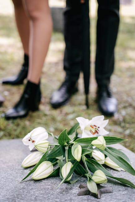 A bouquet of fresh white lilies with open blooms revealing orange and brown anthers, surrounded by closed lily buds and long, slender green leaves, resting on a stone surface. In the background, a per