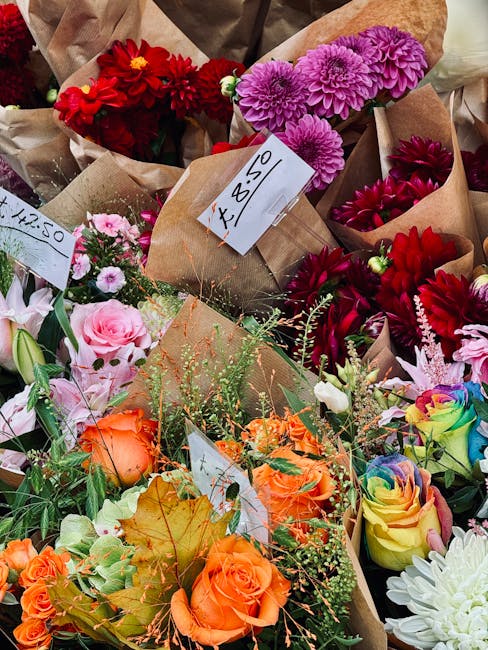 A vibrant collection of freshly cut flowers arranged in brown kraft paper wraps for flower delivery or floral gifting. The display includes red dahlias and deep pink chrysanthemums at the top, with th