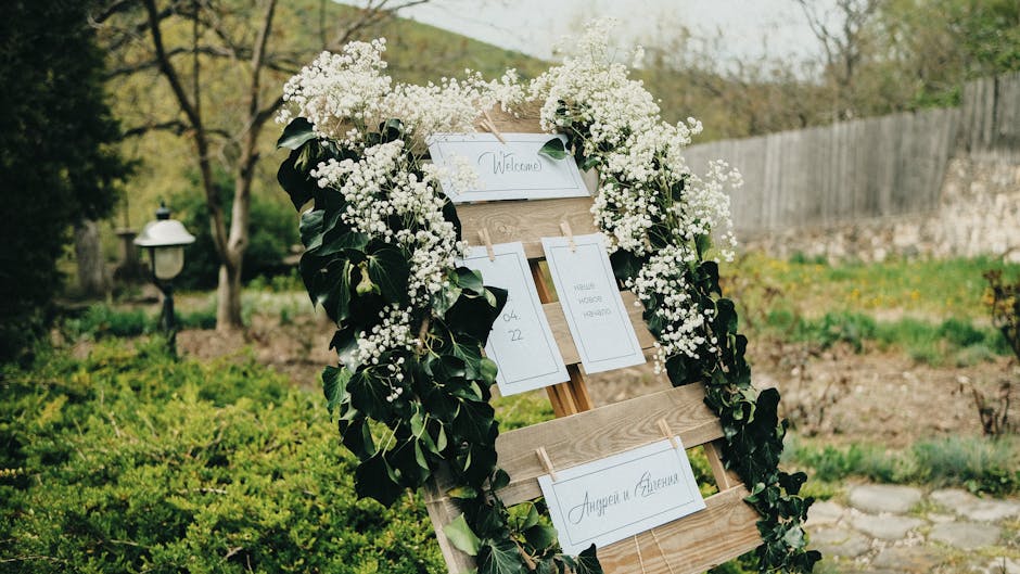 A wedding welcome board decorated with a lush arrangement of white gypsophila and green foliage, mounted on a wooden easel outdoors. The display features several white cards with handwritten calligrap