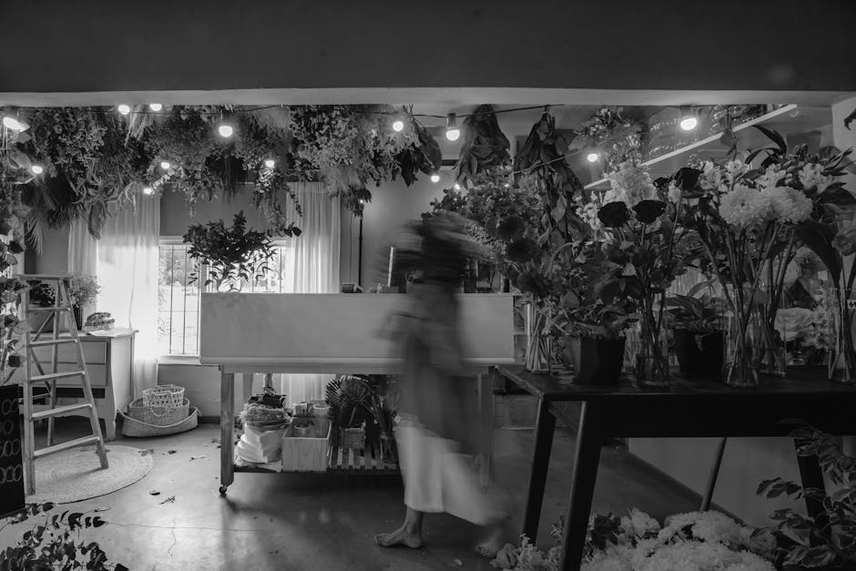 A florist in a workshop capturing the scene with a blurred motion of a woman in a light-colored dress, surrounded by an abundant display of fresh flowers in various stages of arrangement. The space fe