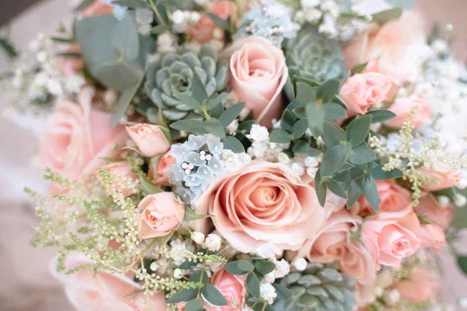 A close-up of a floral arrangement comprising pale pink roses, light blue succulent plants, small white baby's breath, and dusty miller foliage with silvery-grey leaves. The flowers are freshly cut, w