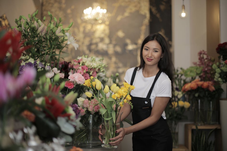 A young woman with long dark hair, dressed in a white t-shirt and black apron, is arranging a bouquet of yellow tulips and light pink roses in a clear glass vase at a floristry shop. The background fe