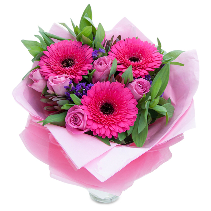 There is a playful elegance to this hand-tied bouquet: three charismatic Gerbera daisies in a saturated fuchsia dominate the view, their concentric petals forming a bold, joyful focal point around dark, almost black centres. Surrounding them, softly muted lavender-pink roses begin to unfurl, each petal layered with care and presenting a velvety texture that absorbs and reflects light in warm, flattering tones. Scattered throughout are small clusters of purple statice, their papery florets adding sprightly punctuation and a cooler contrast to the warm pink family. The greenery is generous - wide, glossy leaves that could be salal paired with the cooler tones of eucalyptus - giving a natural fullness and a slightly dewy visual freshness that suggests a morning harvest. The bouquet is wrapped with intention: an inner pale pink tissue creates a comforting base while an outer translucent, crinkled fuchsia-pink cellophane lends a subtle sheen and modern finish. At the lower edge the top rim of a clear glass vase is visible, the simple transparency of the glass grounding the arrangement and making it immediate and display-ready. The overall scent is delicate - the roses offer a faint floral perfume and the gerberas a clean, green note - creating an atmosphere of tender celebration. Perfect for bringing warmth to a dining table, marking a special date, or surprising someone with a thoughtful gesture, this arrangement reads as both intimate and festive, an expression of care that feels handcrafted and sincere.